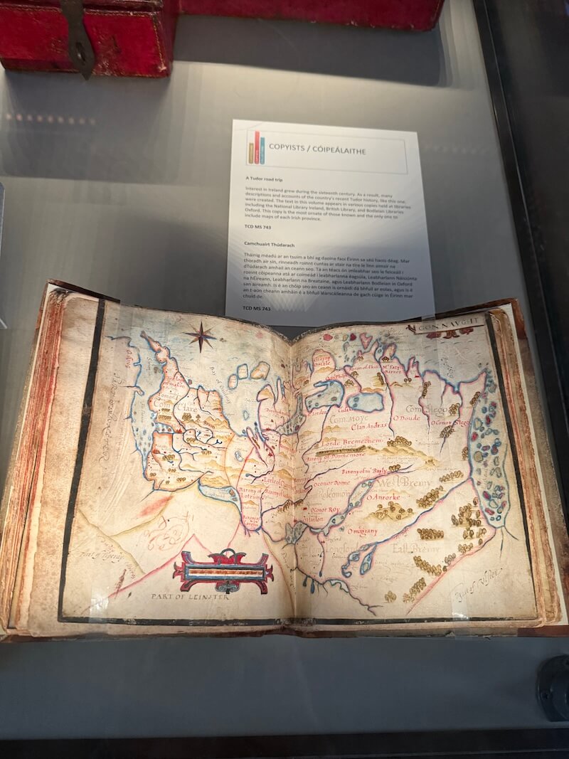 Open manuscript on display under glass featuring a hand-drawn and coloured 16th-century map of Ireland, showing regional names and geographical features, with an information card above titled “Copyists / Cóipeálaithe” describing Tudor-era travel and mapping.