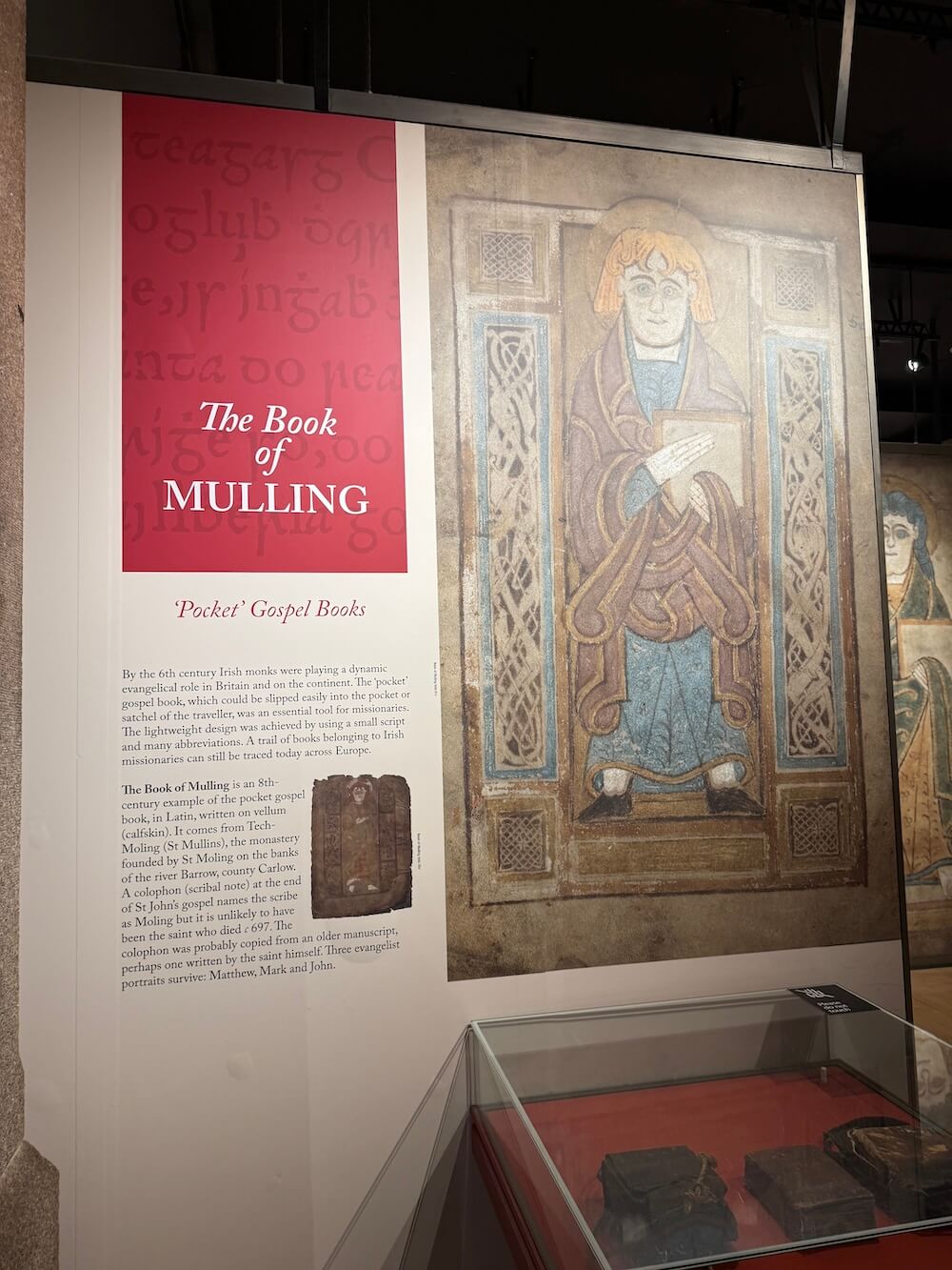 Exhibition display about the Book of Mulling, an 8th-century Irish pocket gospel book, with a large illustrated figure of a saint, explanatory text on its monastic origins, and a glass case containing the ancient manuscript.