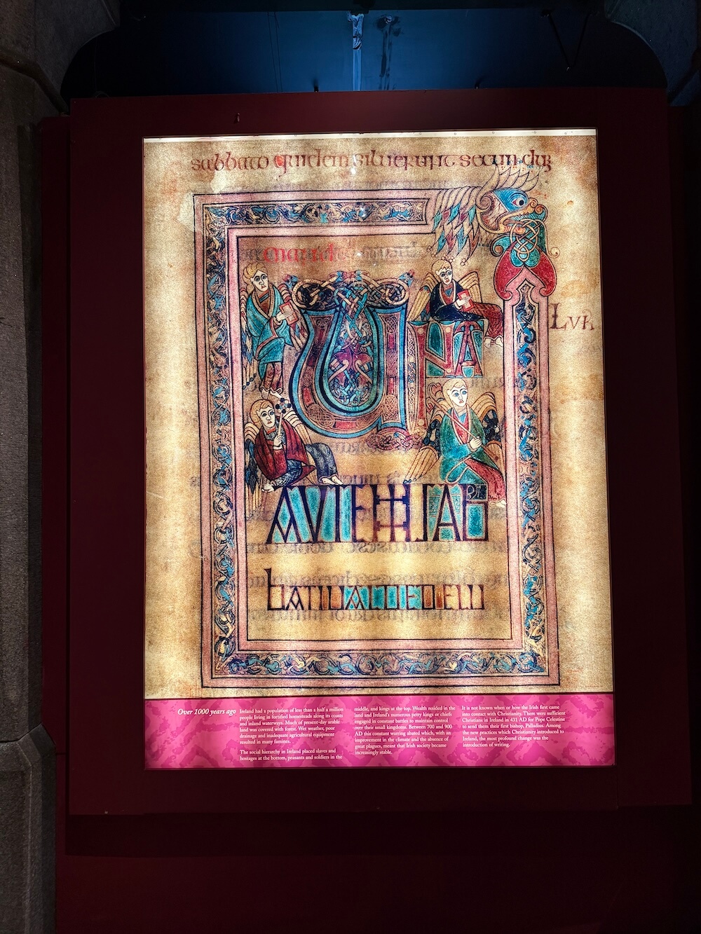 Illuminated manuscript page from the Book of Kells featuring a richly decorated initial letter surrounded by stylised figures and intricate Celtic designs, displayed on a lit panel with red framing.