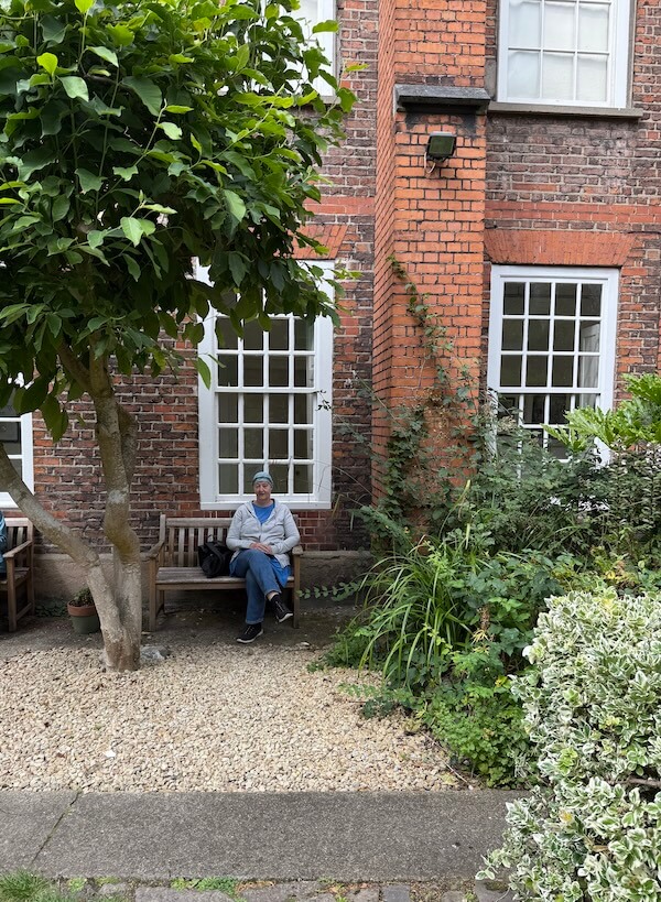 Author sits on a wooden bench in a quiet garden courtyard outside Marsh’s Library, surrounded by brick walls, large sash windows, and leafy greenery under a shady tree.