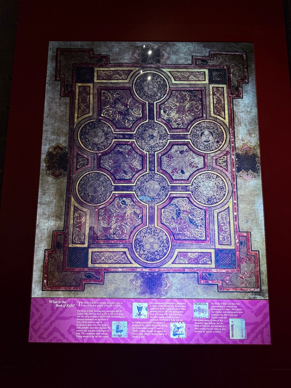 Vibrant illuminated carpet page from the Book of Kells with symmetrical knotwork, symbolic animals, and geometric patterns in red, purple, and gold hues, accompanied by a panel explaining its historical context.