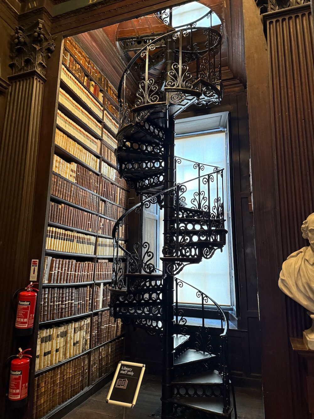 Ornate black wrought-iron spiral staircase leading to the upper level of the Long Room, surrounded by floor-to-ceiling shelves of historic books and a sign marked “Library staff only.”