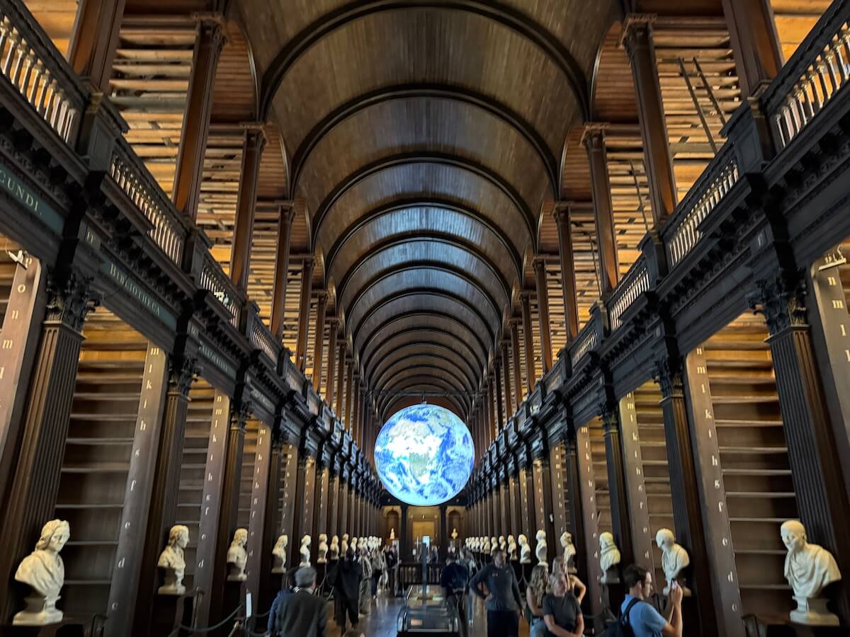 Wide-angle view of the Long Room’s vaulted wooden ceiling and symmetrical rows of marble busts, with a glowing globe of Earth suspended at the far end drawing visitors’ attention.