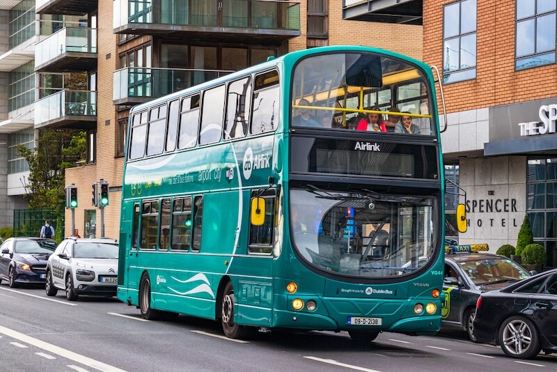 Teal double-decker Airlink bus driving through Dublin city centre, with “Airport City” written on the side and passengers visible on the top deck. The bus is passing The Spencer Hotel and surrounded by cars and modern buildings.
