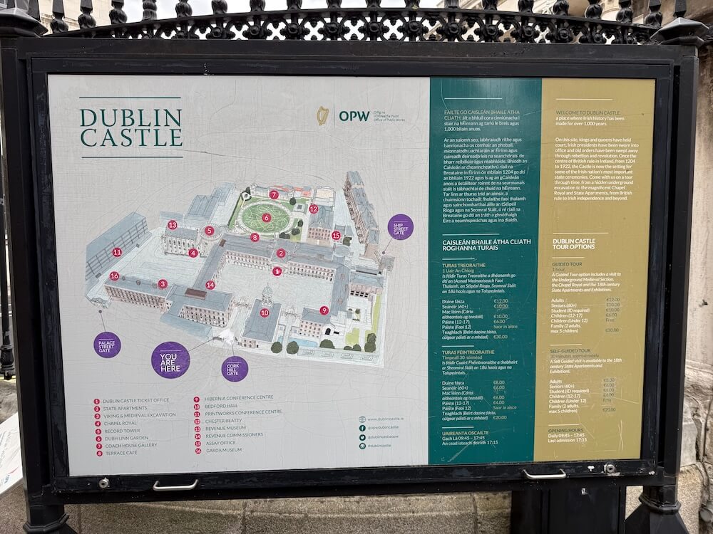 Outdoor visitor information board for Dublin Castle featuring a detailed site map with numbered landmarks such as the State Apartments, Chapel Royal, and Dubh Linn Garden. Text sections provide historical background in both English and Irish, as well as details about guided and self-guided tour options and opening hours