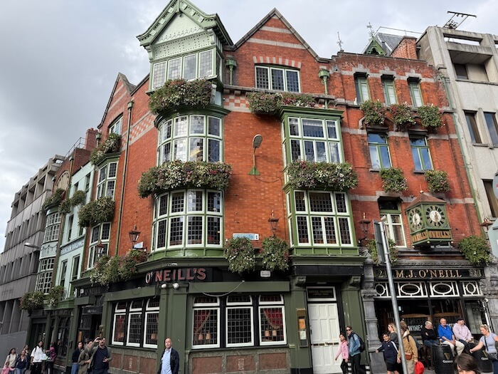 Historic red-brick pub building on a street corner in Dublin, identified as O’Neill’s, with bay windows framed in pale green and flower boxes overflowing with greenery and blooms