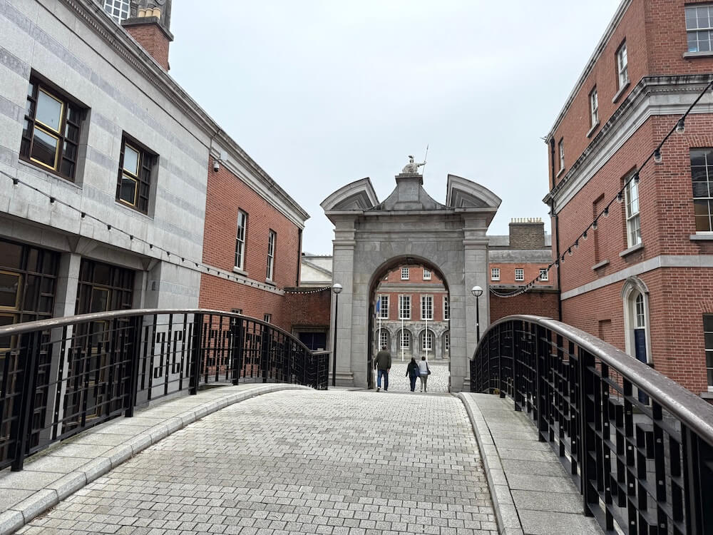 Arched stone gateway at Dublin Castle viewed from a gently curving pedestrian bridge, flanked by red-brick and grey-stone buildings. Three people walk beneath the arch toward a courtyard lined with symmetrical Georgian windows.