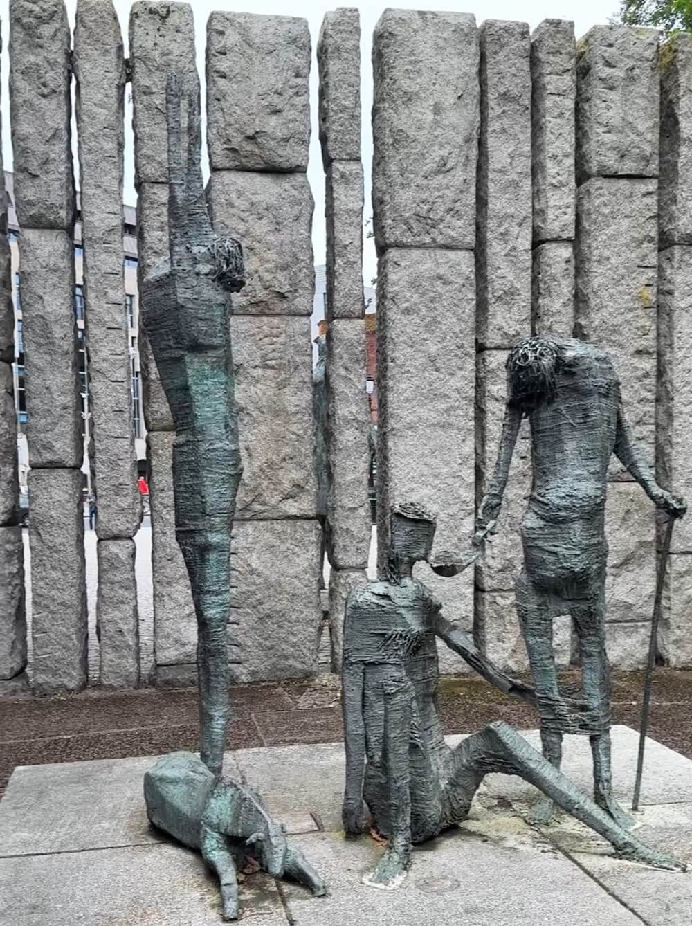Modern bronze sculpture of thin, abstract human figures and a dog, part of the Famine Memorial in Dublin. The emaciated forms are positioned against a backdrop of tall stone slabs, evoking the suffering of the Great Irish Famine.