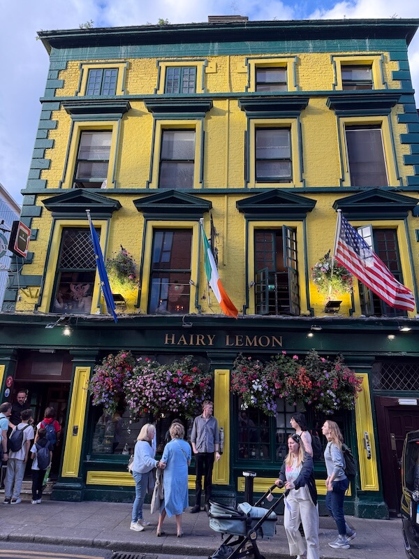 Vibrant yellow and green facade of The Hairy Lemon pub, decorated with hanging flower baskets and flying the Irish, EU, and American flags, with groups of people gathered outside.