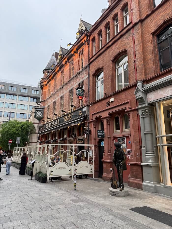 Brick-fronted Bewley’s Grafton Street café and bar with ornate detailing and a patio area, next to a bronze statue of Phil Lynott holding a guitar, standing on a cobbled pedestrian street in Dublin.