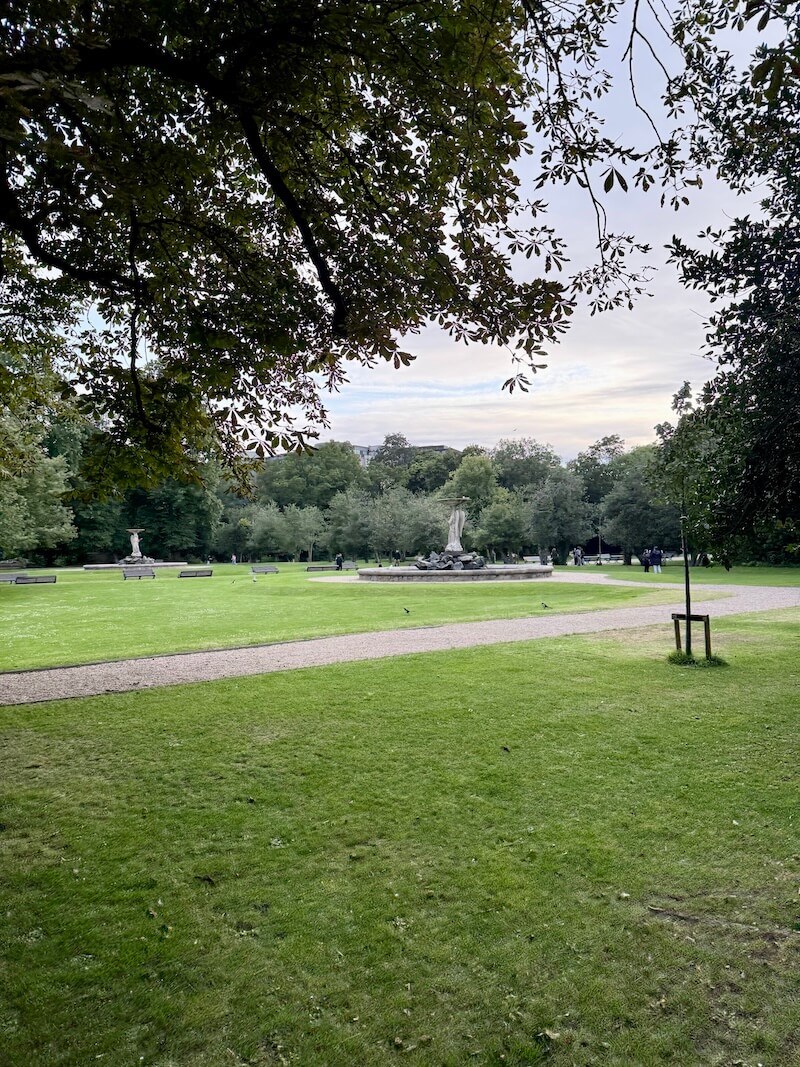 A wide grassy lawn in St. Stephen’s Green, Dublin, with a central fountain surrounded by benches and trees. The early evening sky is visible through the overhanging tree branches in the foreground.