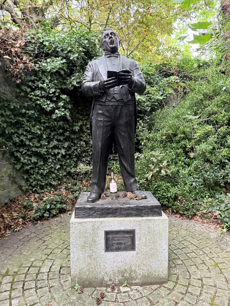 This is a statue of Irish tenor John McCormack, located in Dublin’s Iveagh Gardens. He’s shown mid-performance, holding sheet music, surrounded by pinecones—and apparently a fan left a bottle too!