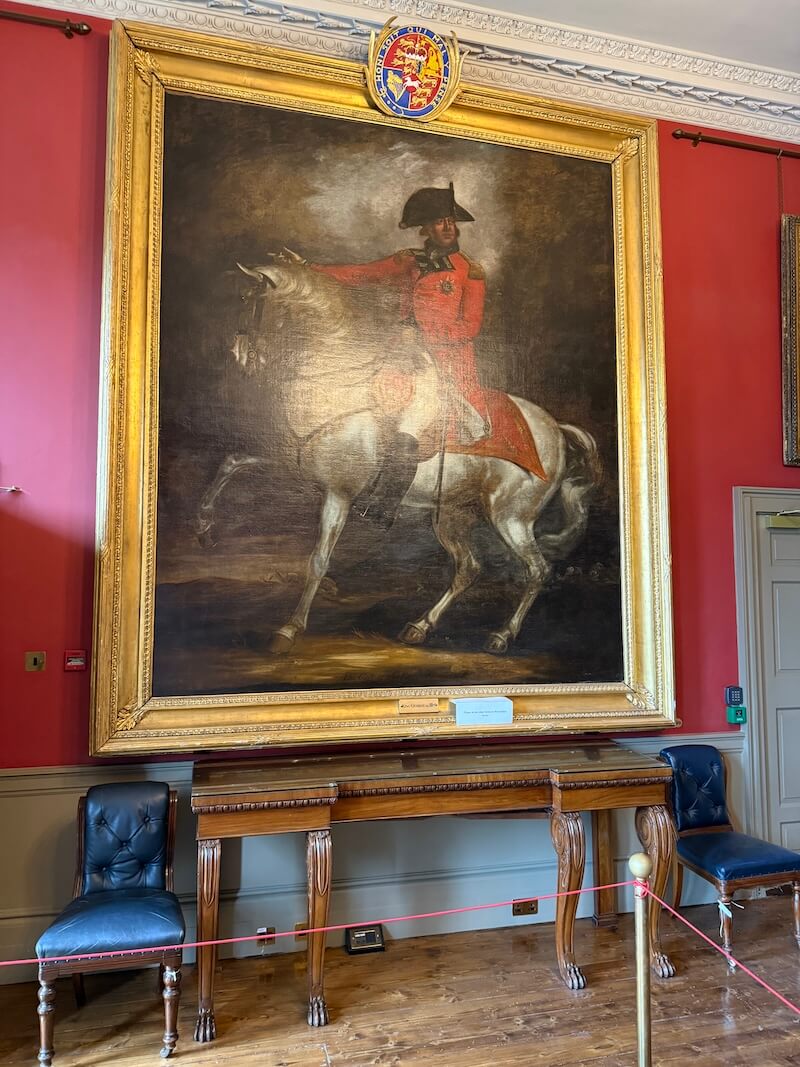 Large equestrian portrait of a military figure in a red coat and bicorne hat, displayed in a grand frame above a wooden table and flanked by two navy leather chairs in an opulent red-walled room.