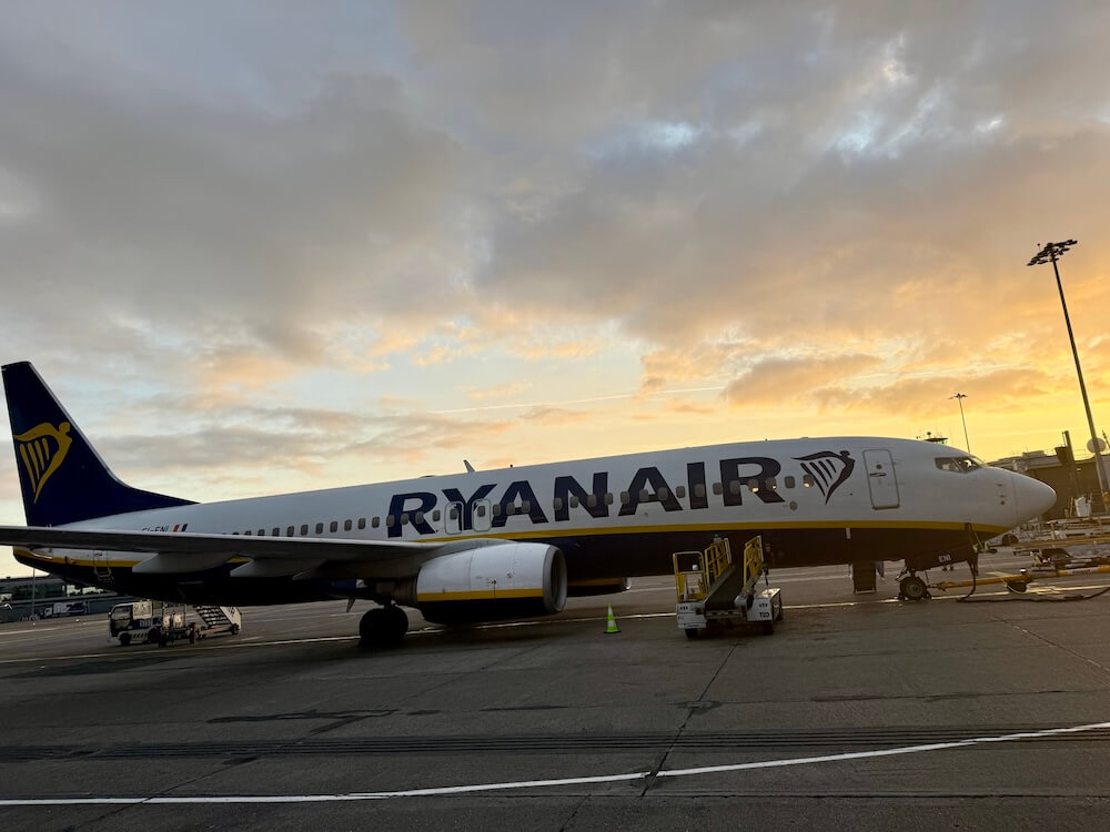 Ryanair aircraft parked on the tarmac at sunset, with golden light reflecting off the fuselage and dramatic clouds in the background. Ground service vehicles and boarding stairs are positioned beside the plane.