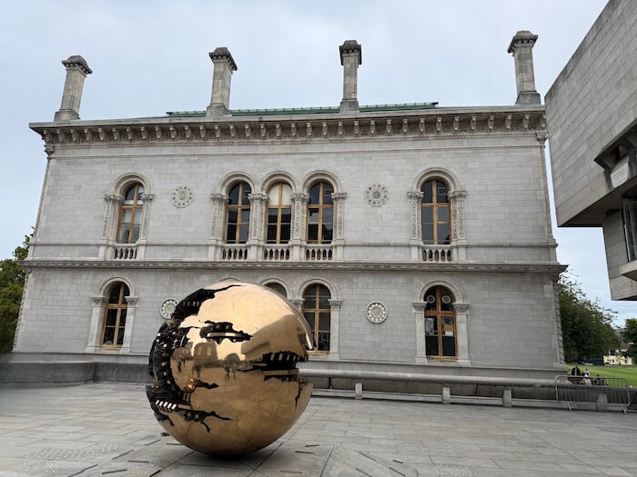 The bronze "Sphere Within Sphere" sculpture by Arnaldo Pomodoro displayed in front of a grand neoclassical building at Trinity College Dublin, with arched windows and decorative columns.