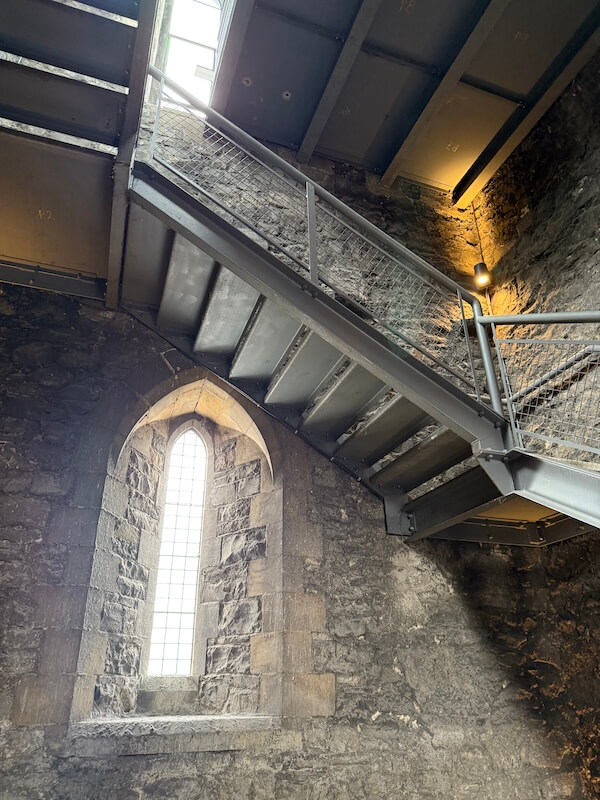 The interior of St. Michael’s Tower at Dublinia. The modern metal stairs lead to the observation deck, with a tall Gothic window highlighting the historic stone walls.