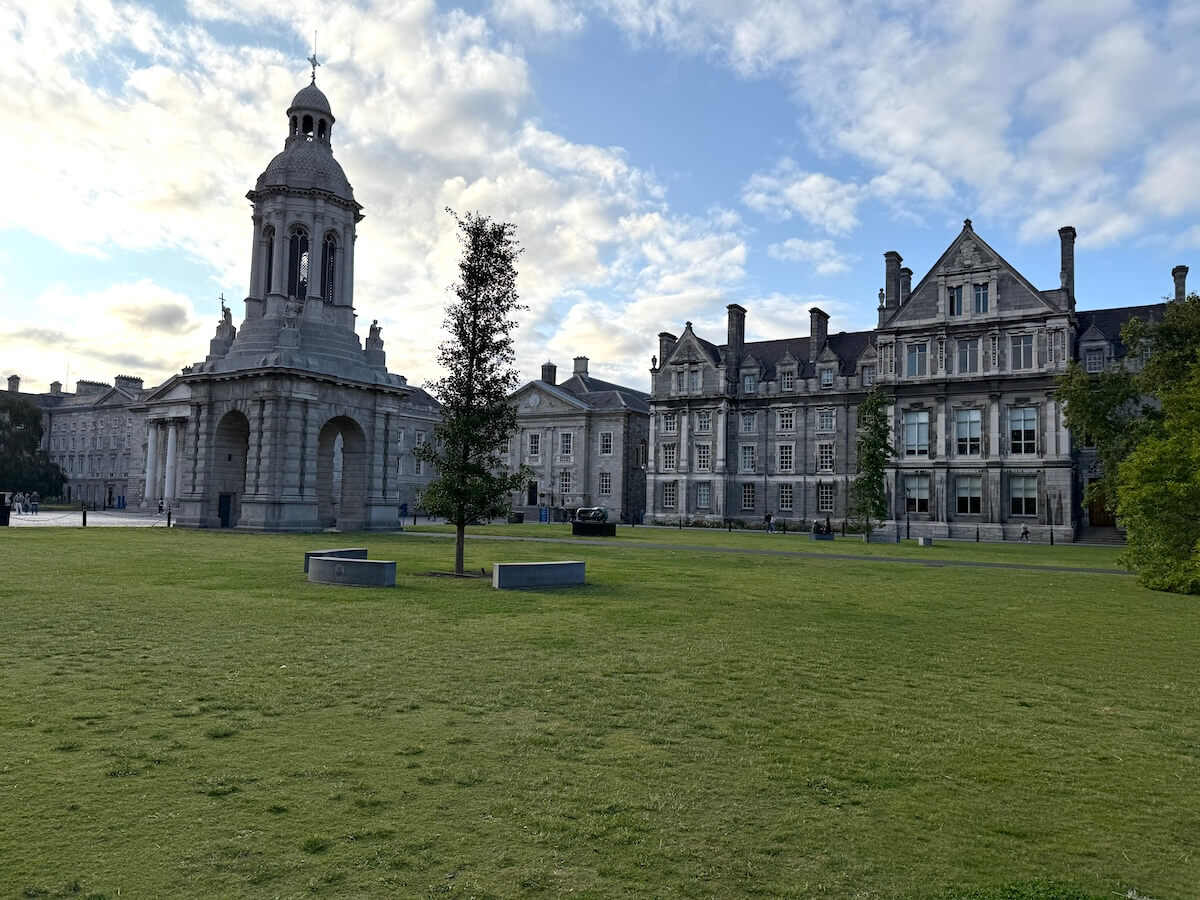 Wide-angle view of the Campanile bell tower and surrounding classical buildings in the main quad of Trinity College Dublin, with a green lawn and a few modern benches in the foreground under a partly cloudy sky.