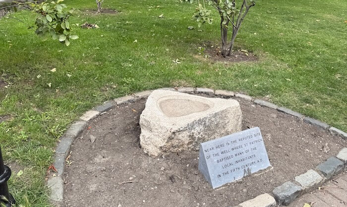 Triangular stone basin set in a circular patch of soil with a plaque that reads, “Near here is the reputed site of the well where St Patrick baptized many of the local inhabitants in the fifth century A.D.” The site is surrounded by neatly maintained grass and small trees.