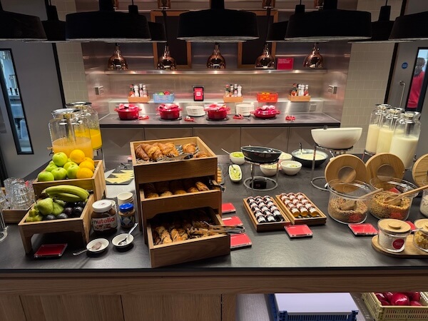 Well-lit hotel breakfast buffet with a variety of items including fresh fruit, pastries, cereals, glass bottles of milk, fruit juices, and spreads like Nutella. The setup includes wooden crates and dispensers, with hot food in red pots on a counter in the background under copper pendant lights
