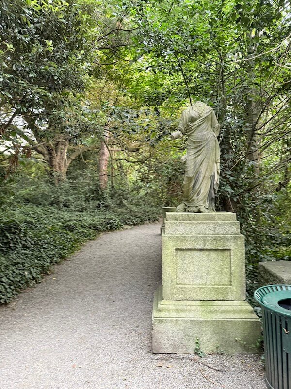 A headless classical-style statue stands beside a quiet gravel path surrounded by dense greenery in Iveagh Gardens, lending a mysterious and aged charm to the scene.
