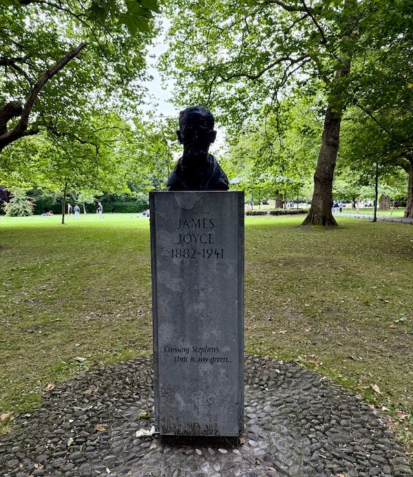 Bronze bust of James Joyce on a tall stone pedestal in St. Stephen’s Green, Dublin. The pedestal is engraved with his name, lifespan (1882–1941), and the quote: “Crossing Stephen’s, that is my green...” surrounded by leafy trees and grass.