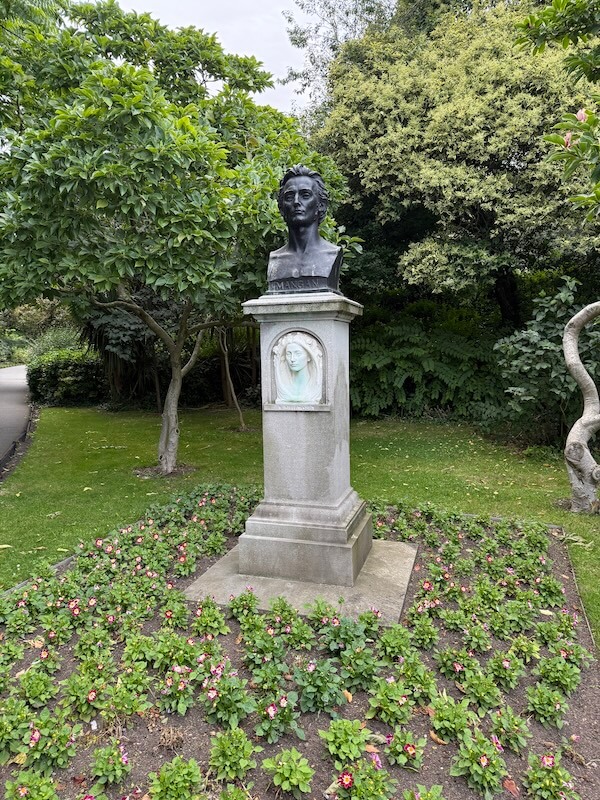 Statue of poet James Clarence Mangan set in a flowerbed, surrounded by green trees and plants. The bust sits atop a stone pedestal with a sculpted relief of a woman beneath the name “J. MANGAN.”