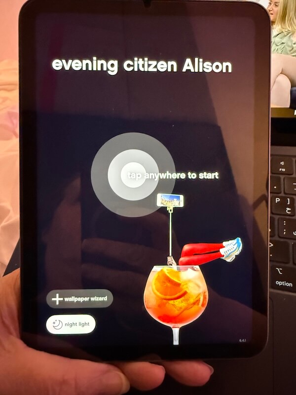 A person holds a tablet displaying a welcome screen that reads “evening citizen Alison,” with a circular button labeled “tap anywhere to start” and a playful graphic of a cocktail with legs sticking out holding a selfie stick.