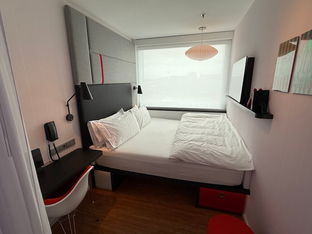 Compact hotel room with a large bed pushed against the window, featuring crisp white bedding and a modern padded headboard. The room includes a small black desk with a red and white chair, wall-mounted lamps, a flat-screen TV, and minimalistic decor with bright accents.