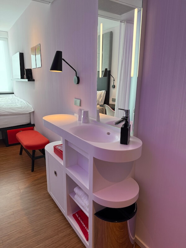 Modern hotel room vanity area with a curved white sink, black soap dispenser, mirror with built-in lighting, and a red bench in front of a made bed. White towels and a red item are neatly stored on the open shelves below the sink.