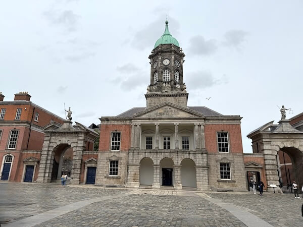 View of the historic Bedford Tower in the Upper Yard of Dublin Castle, featuring a central clock tower with a green dome, red-brick wings, and statues atop arched gateways on either side.