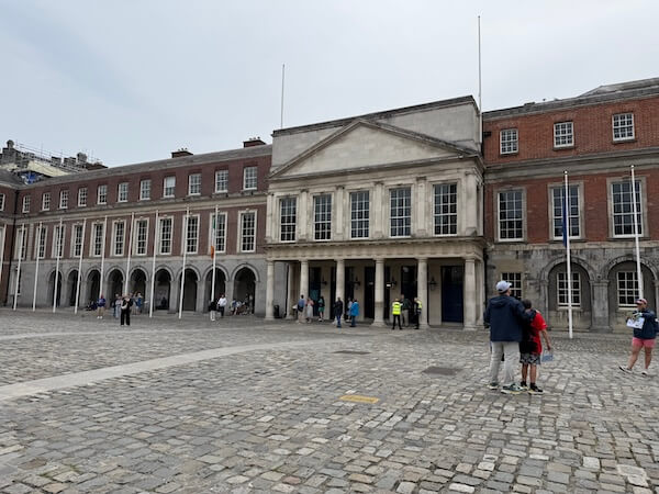 Cobblestone courtyard at Dublin Castle showing the State Apartments building, with a neoclassical façade, arched colonnade, and groups of visitors gathered near the entrance.