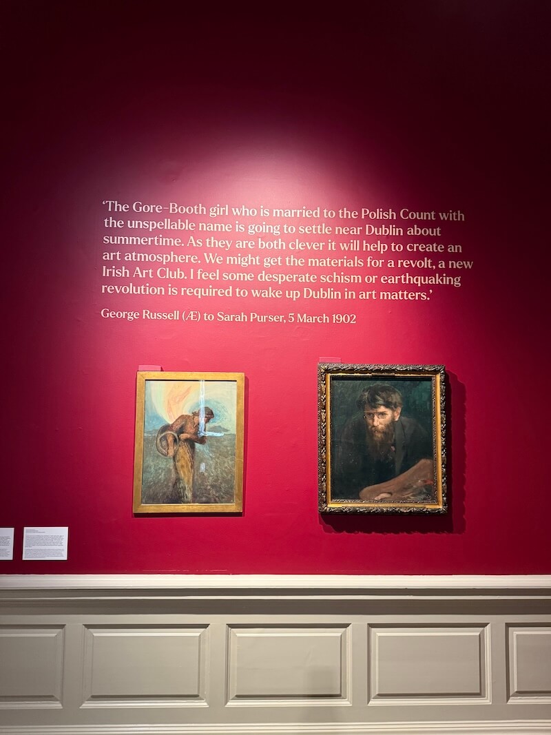 Art gallery wall painted deep red with two framed paintings and a quote from George Russell to Sarah Purser dated 5 March 1902, discussing the arrival of Constance Gore-Booth and her husband near Dublin and their potential to inspire an art revolution.