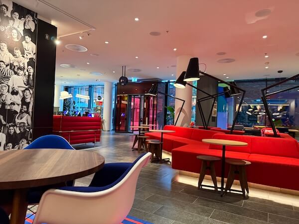 Stylish hotel lounge area with red sofas, round tables, and modern oversized lamps, set against a backdrop of floor-to-ceiling windows and a black-and-white photo mural.