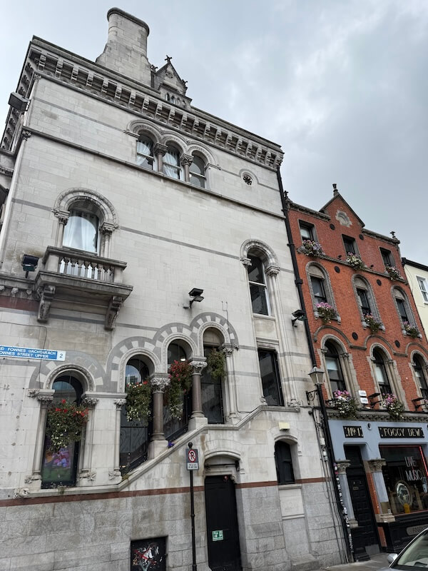 Ornate grey stone building with arched windows and decorative columns in Dublin’s Temple Bar area, next to a red-brick pub with hanging flower baskets and a sign for The Foggy Dew.