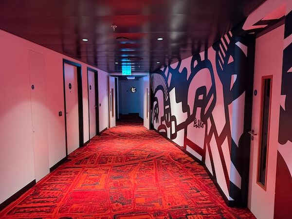 Colourfully lit hotel hallway with a bold red carpet and black-and-white abstract wall art, leading to several guest room doors.