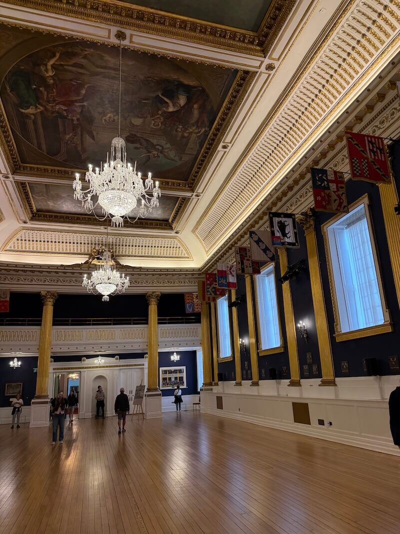 Grand ballroom with a polished wooden floor, ornate ceiling paintings, crystal chandeliers, and tall gold columns, with heraldic banners lining the upper walls and visitors exploring the space.