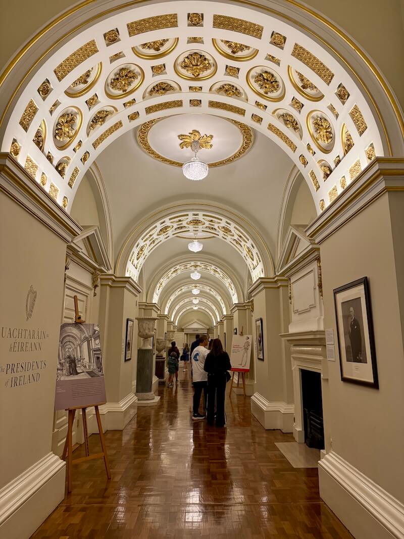 Ornate interior hallway with arched ceilings adorned with gilded rosettes and chandeliers, part of a museum or government building featuring the “Presidents of Ireland” exhibit, with visitors walking through the corridor.
