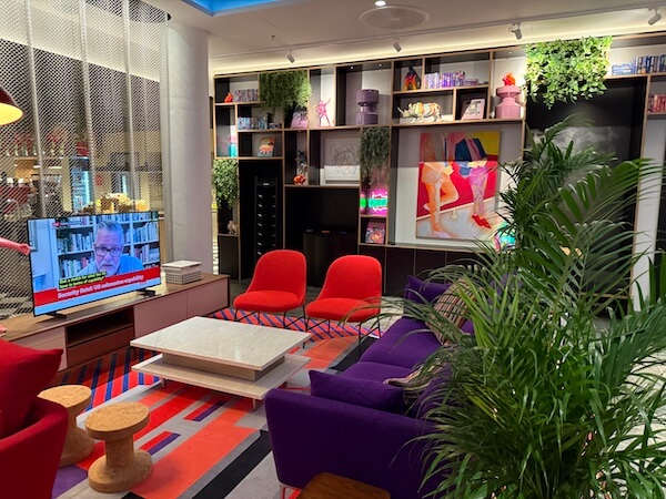 Cosy hotel lounge with vibrant seating including purple and red chairs, a patterned rug, potted plants, and a large TV showing a news segment beside a modern bookshelf wall.