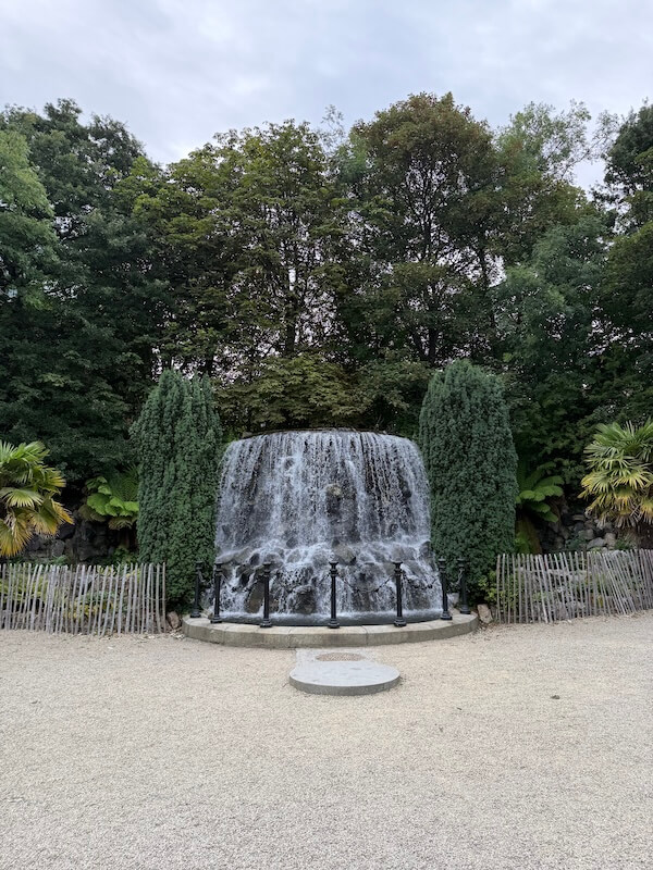 Semi-circular waterfall framed by evergreen trees and wooden fencing in Iveagh Gardens, cascading into a shallow pool bordered by a gravel area.