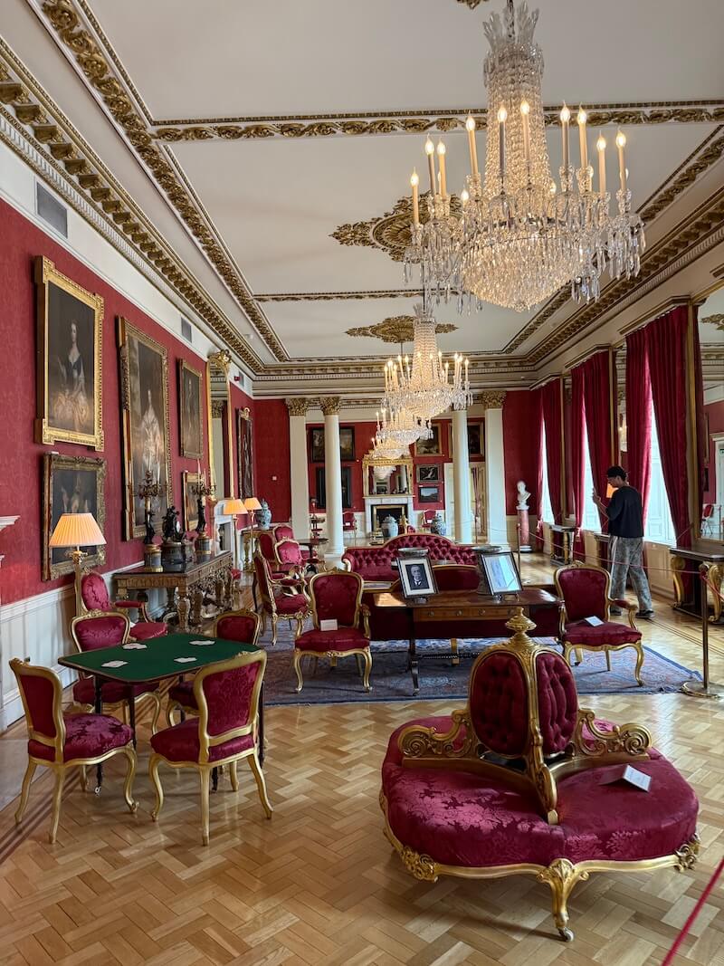 Luxurious red drawing room with velvet furniture, gilt frames, crystal chandeliers, and historic portraits on red damask walls, exuding 18th-century aristocratic elegance.