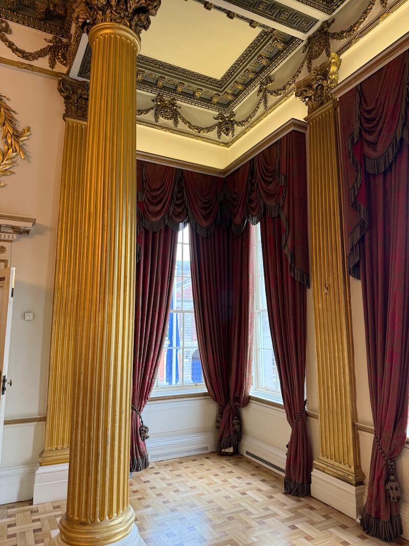 Close-up of a corner in an opulent historic room with large gold fluted columns, intricate ceiling mouldings, and heavy red draped curtains framing tall sash windows.