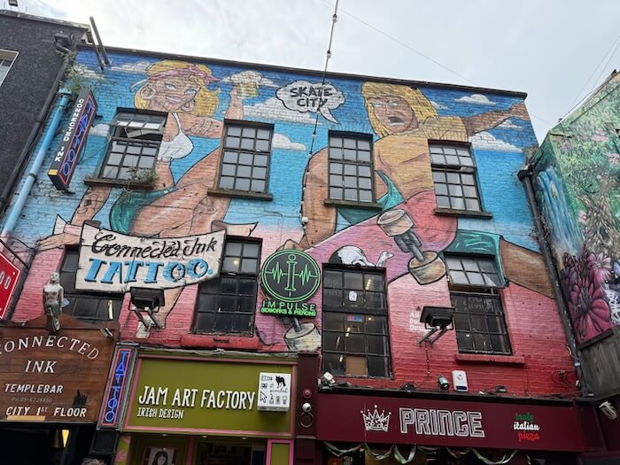 Colourful street mural in Temple Bar, Dublin, featuring cartoon-style roller skaters painted across the upper facade of a building housing tattoo shops, a gym, and small local businesses like Jam Art Factory.