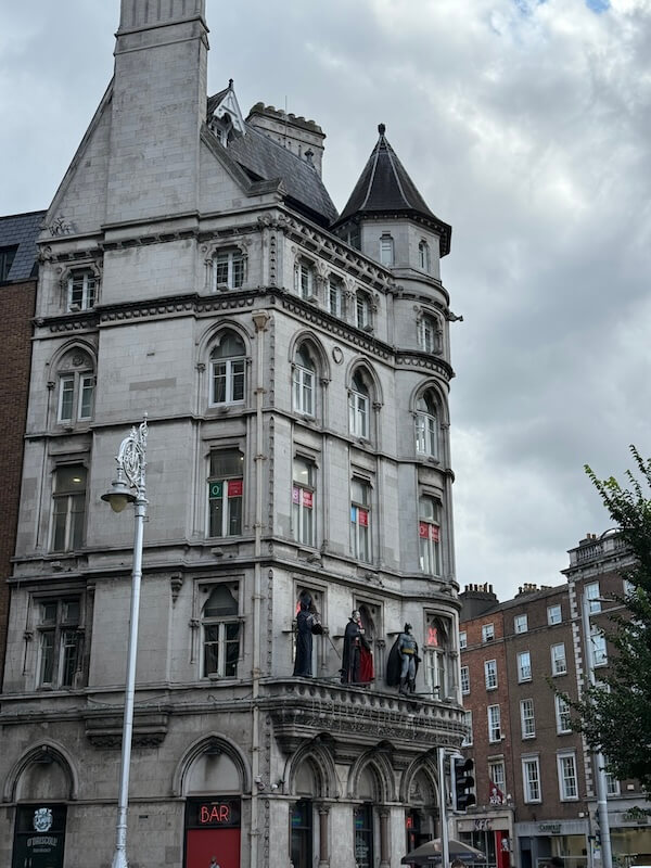 Gothic-style corner building in Dublin with turrets and stone statues of cloaked figures on a balcony, contrasting sharply against the modern signage for a bar below.
